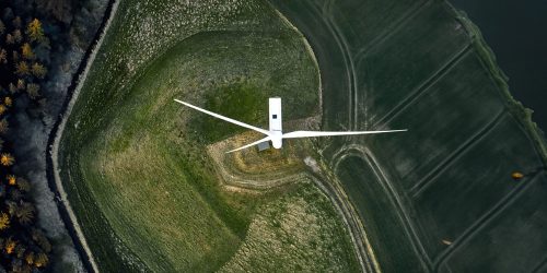 Arial view of a solitary wind turbine in a lush green field bordering a dense forest, highlighting renewable energy.