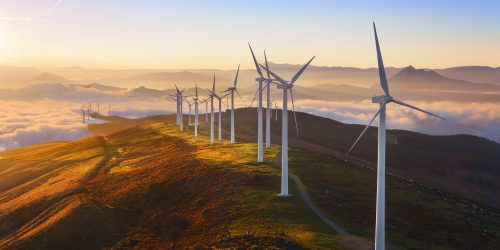 Sunrise over wind farm on a hill with turbines turning, set against a backdrop of clouds and mountains. Renewable energy landscape.